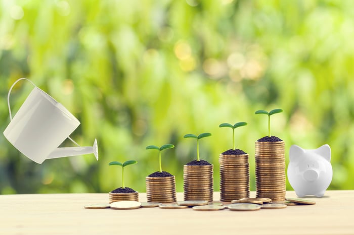 Rending of a watering can next to increasingly taller stacks of coins with plants sprouting from each stack next to a piggy bank, illustrating the concept of compound interest. 